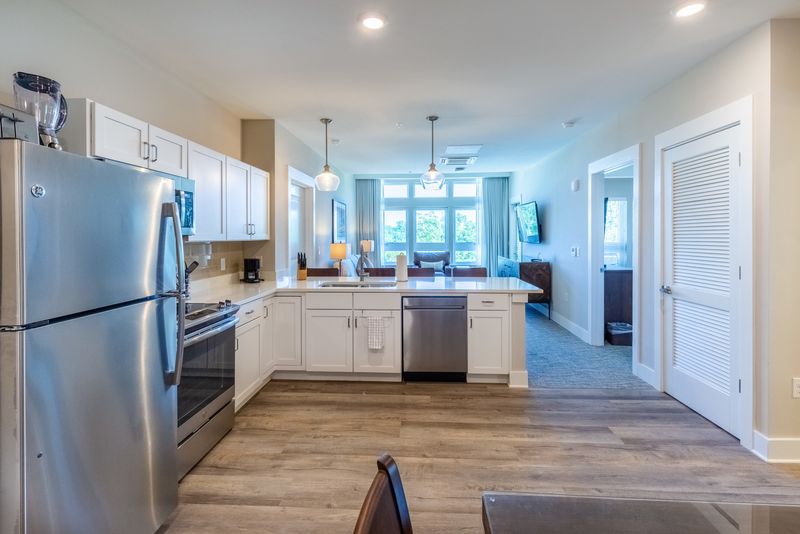 Silver appliances, white cabinets, and white countertops adorn the kitchen, while a large window baths the living area in natural light.